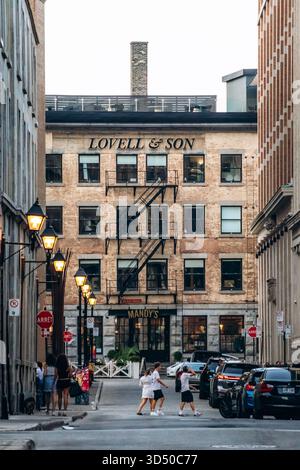 Montreal, Kanada - 11. August 2025: Abendliche Stadtlandschaft mit historischem Lovell und Son Gebäude, Vintage Straßenlaternen, Autos und Menschen in der Rue le Moyne in Stockfoto