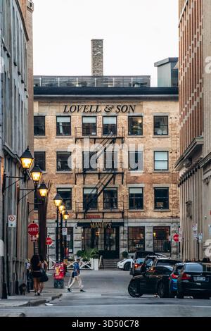 Montreal, Kanada - 11. August 2025: Abendliche Stadtlandschaft mit historischem Lovell und Son Gebäude, Vintage Straßenlaternen, Autos und Menschen in der Rue le Moyne in Stockfoto