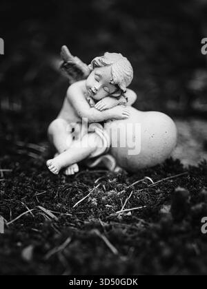 Schwarz-weiße Nahaufnahme einer schlafenden Engelsstatue auf einem Herzen auf einem Friedhof. Stockfoto