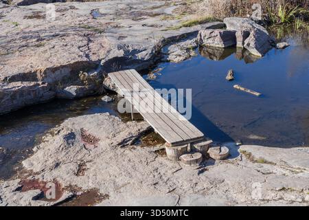 Holzsteg über flachen, felsigen Bach mit ruhigem, klarem Wasser und Steinlandschaft an einem sonnigen Tag im Freien Stockfoto