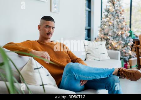 Afroamerikaner sitzt zu Hause auf dem Sofa und trägt einen orangefarbenen Pullover neben dem Weihnachtsbaum Stockfoto