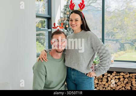 Ein Paar posiert und lächelt am Weihnachtsbaum und Brennholz im Wohnzimmer mit Geweih-Haarbändern Stockfoto