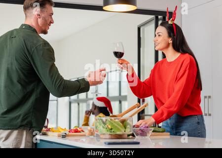 Weißes Paar hebt Weingläser auf der Kücheninsel, afroamerikanischer Mann hackt Gemüse Stockfoto
