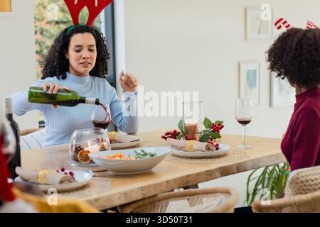 Verschiedene Freundinnen in Geweih, die Wein am Esstisch mit Weingläsern und Kopierraum gießen Stockfoto