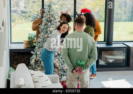 Verschiedene Freunde schmücken den beflockten Weihnachtsbaum im Wohnzimmer und tauschen eingepackte Geschenke aus Stockfoto