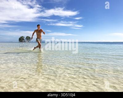 Ein junger, sportlicher, gutaussehender Mann, der durch das flache Meer an einem sonnigen Strand läuft. Das sportliche männliche Modell zeigt einen aktiven Lebensstil, Stärke. Stockfoto