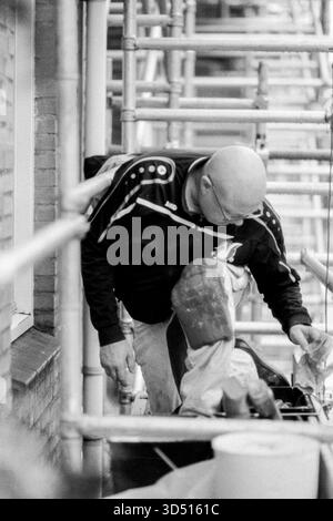 Maler, der an Gerüsten arbeitet. Industriemalerei und Bauarbeiter, arbeitet an einem Hausjob in Immobilien von einem dreistöckigen Gerüstbau aus. Tilburg, Niederlande. Aufgenommen mit analogem Schwarzweißfilm, 2022. Tilburg Studio Tuinstraat Noord-Brabant Nederland Copyright: XGuidoxKoppesxPhotox Stockfoto