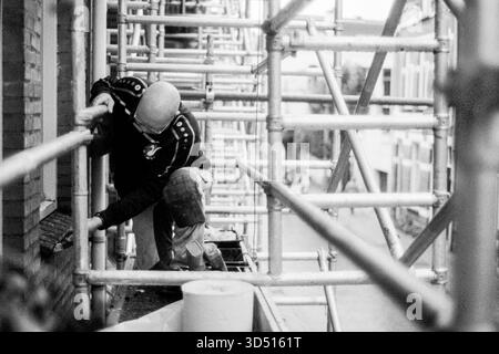 Maler, der an Gerüsten arbeitet. Industriemalerei und Bauarbeiter, arbeitet an einem Hausjob in Immobilien von einem dreistöckigen Gerüstbau aus. Tilburg, Niederlande. Aufgenommen mit analogem Schwarzweißfilm, 2022. Tilburg Studio Tuinstraat Noord-Brabant Nederland Copyright: XGuidoxKoppesxPhotox Stockfoto