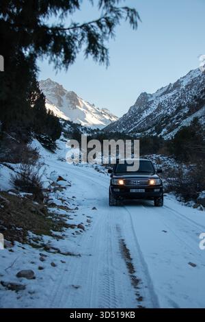 Naryn, Kirgisistan – 13. Dezember 2024: Ein schwarzer Toyota Land Cruiser 100 mit Scheinwerfern auf einer schneebedeckten Straße durch ein Tal mit Dramatten Stockfoto
