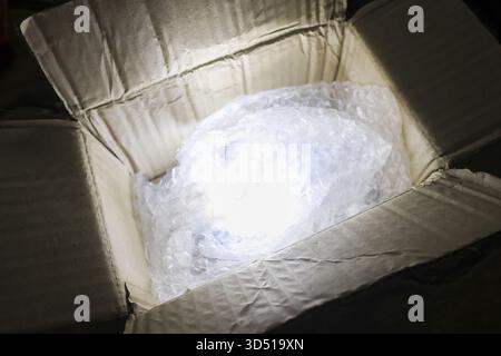 Geheimnisvolle offene Kartonbox, die von innen mit hellem Licht leuchtet. Zerbrechliches Paket in Luftpolsterfolie sorgt für Überraschung und Entdeckung Stockfoto