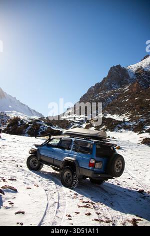 Naryn, Kirgisistan - 12. November 2025: Ein stark modifizierter blauer Geländewagen mit großen Reifen und Dachkoffer fährt auf tiefem Schnee in einem schneebedeckten, schneebedeckten Mo Stockfoto