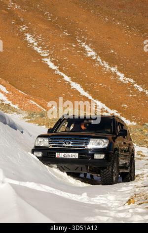 Naryn, Kirgisistan – 12. November 2025: Ein schwarzer Toyota Land Cruiser 100 fährt auf einem verschneiten Bergweg, eingerahmt von einem steilen, rötlich-braunen trockenen Hügel Stockfoto