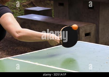 Die fokussierte Hand hält ein schwarzes Paddel und spielt im Freien Tischtennis. Ein orangener Ball schlägt Schläger während sportlicher Aktivitäten auf dem grünen Tisch in PA Stockfoto