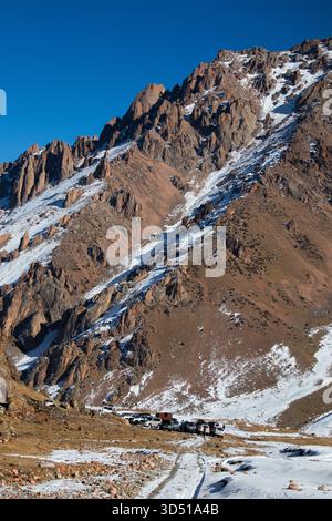 Naryn, Kirgisistan – 12. November 2025: Eine Linie von Geländewagen (SUV-Konvoi) parkte auf einer unbefestigten und schneebedeckten Piste am Fuße eines massiven, zerklüfteten, rötlichen B Stockfoto