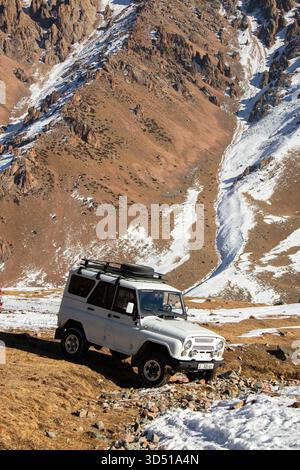 Naryn, Kirgisistan – 12. November 2025: Ein weißer Patriot der UAZ parkt auf einem schneebedeckten Hügel gegen einen massiven, steil abfallenden, rötlich-braunen Berg Stockfoto