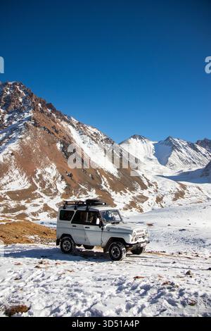 Naryn, Kirgisistan - 12. November 2025: Ein weißer Patriot der UAZ, der auf einer verschneiten Ebene parkt und einem dramatischen Tal aus schneebedecktem und rötlich-braunem Moun gegenübersteht Stockfoto