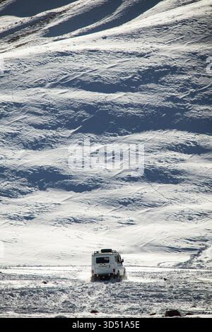 Naryn, Kirgisistan - 12. November 2025: Ein weißer UAZ Patriot 4x4 fährt über eine verschneite Ebene in Richtung Kamera, mit einem riesigen, strukturierten schneebedeckten Hügel Stockfoto