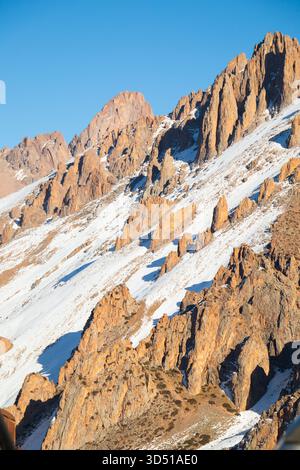 Dramatische, steile, felsige Berghänge mit orange-braunen Erdtönen und kontrastierenden vertikalen Schneeflächen bei klarem Sonnenlicht Stockfoto