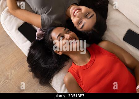 Lachende Freundinnen, die zusammen auf der Couch zu Hause liegen, mit Smartphones neben den Köpfen Stockfoto