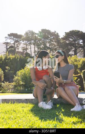 Unterhalten Sie sich mit verschiedenen Freundinnen, die auf einem Steinvorsprung im sonnigen Park sitzen, mit Smartphone Stockfoto