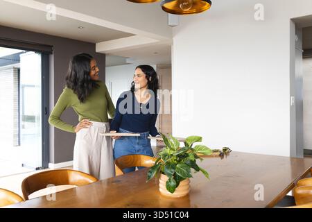 Verschiedene Freundinnen stehen am Holztisch im Esszimmer, halten eine Diele und ein Maßband Stockfoto