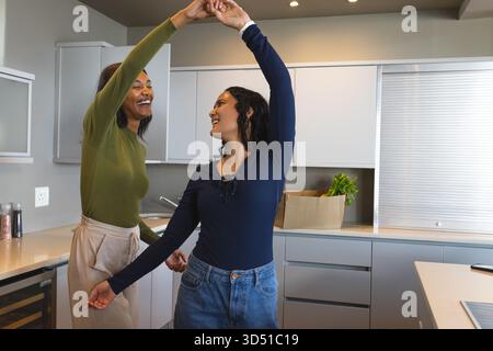 Verschiedene Freundinnen, die sich in einer modernen Küche drehen und tanzen, mit Pappschachtel mit Lebensmitteln Stockfoto