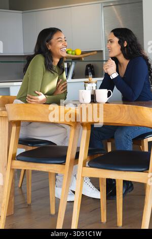 Lachen und plaudern verschiedene Freundinnen, die am Küchentisch sitzen, mit Kaffeetassen Stockfoto