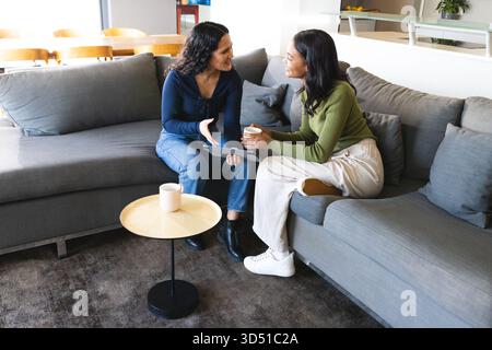 Unterhalten Sie sich mit verschiedenen Freundinnen, die auf einem grauen Sofa im Wohnzimmer sitzen, mit Tablet und Kaffeetassen Stockfoto