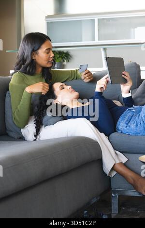 Stöbern Sie in verschiedenen weiblichen Paaren mit Tablet auf grauem Sofa im modernen Wohnzimmer, mit Kreditkarte Stockfoto