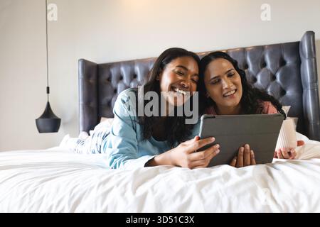 Liegend verschiedene Freundinnen, die sich ein Tablet auf dem Bett teilen, im modernen Schlafzimmer, mit Keramikbecher Stockfoto