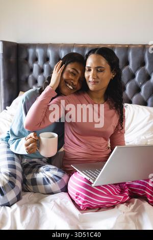 Verschiedene Partnerinnen kuscheln, die auf dem Bett im Schlafzimmer einen silbernen Laptop suchen, mit weißem Keramikbecher Stockfoto