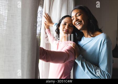 Verschiedene Frauen, die den Vorhang zurückziehen und zu Hause am Fenster sanft lächeln, mit transparenten Vorhängen Stockfoto