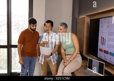 Verschiedene Kollegen, die gedruckte Dokumente mit Markierungen im modernen Büro mit offenem Laptop überprüfen Stockfoto