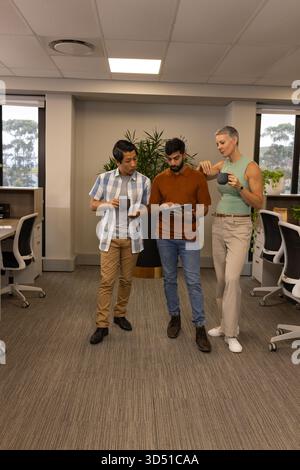 Verschiedene Kollegen stehen, die Inhalte auf einem Tablet im Bürokorridor mit Kaffeetassen überprüfen Stockfoto