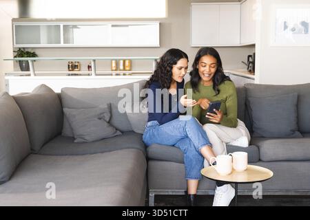 Lächelnde, vielseitige Freundinnen, die sich auf einem grauen Sofa im Wohnbereich mit Smartphone und Keramikbecher unterhalten Stockfoto
