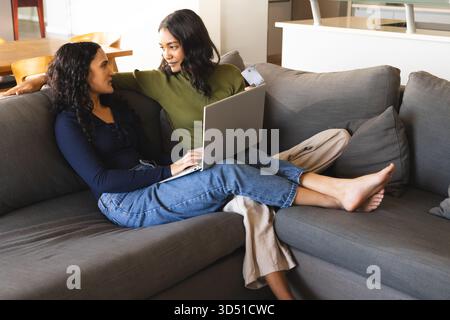 Entspannen Sie verschiedene weibliche Paare online auf Laptop in einem modernen Wohnzimmer, mit Kreditkarte Stockfoto