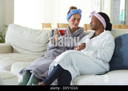 Entspannende, vielfältige Freundinnen, die sich auf dem Sofa im Wohnzimmer unterhalten, mit weichen Bademänteln und Teetassen Stockfoto