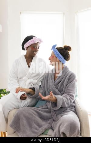 Verschiedene Freundinnen sitzen im Wohnzimmer auf einem Sessel, mit Bademänteln und Patches unter den Augen Stockfoto