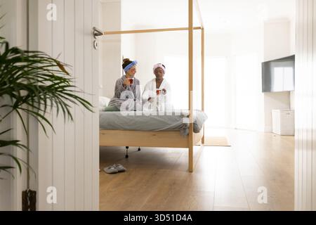 Entspannende Freundinnen, die auf dem Bett sitzen und klare Tassen im Schlafzimmer halten, mit Himmelbett Stockfoto
