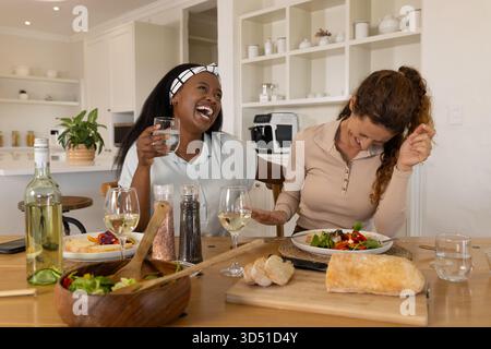 Lachende, vielfältige Freundinnen, die Salat und Wein an einem modernen Esstisch mit Brot teilen Stockfoto