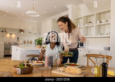 Wein gießen, verschiedene Freundinnen unterhalten sich an einem modernen Küchentisch mit frischem Salat Stockfoto