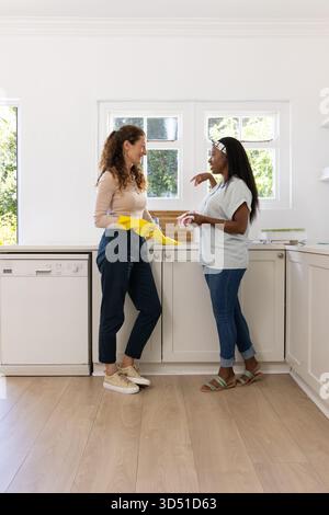 Verschiedene Freundinnen, die Geschirr in der Küche in der Nähe des Geschirrspülers reinigen, gelbe Handschuhe halten Stockfoto