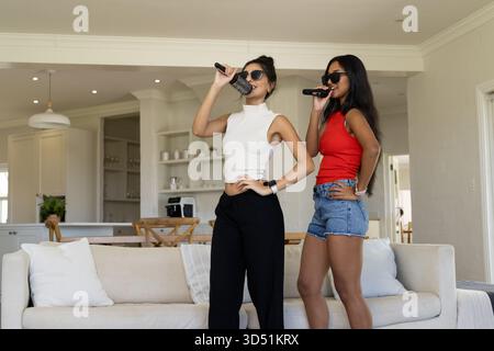 Sie singen verschiedene weibliche Freunde mit Karaoke-Mikrofonen im Wohnzimmer und weißen Sofakissen Stockfoto Sie singen verschiedene weibliche Freunde mit Karaoke-Mikrofonen im Wohnzimmer und weißen Sofakissen Stockfoto