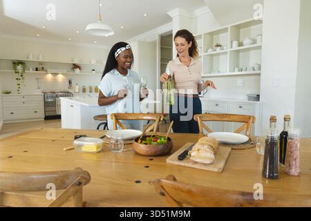 Verschiedene Freundinnen, die Weißwein gießen und leere Gläser in der Küche halten, mit Esstisch Stockfoto