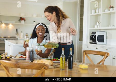 Wein gießen und plaudern, verschiedene Freundinnen sitzen an einem modernen Küchentisch mit Salatplatten Stockfoto