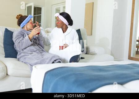 Verwöhnen Sie verschiedene weibliche Freunde, die rote Tassen auf weißem Sofa zu Hause halten, und tragen Sie unter-Augen-Masken auf Stockfoto