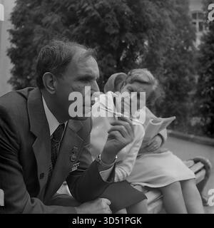 Dieses leistungsstarke Schwarzweiß-Archivfoto aus dem Jahr 1975 ist ein intimes Profilporträt des blinden Veteranen Shutenko in Swiatohirsk (Slavyanogorsk), ukrainische SSR. Während des 30. Siegtages aufgenommen, fängt es einen ruhigen, nachdenklichen Moment ein. Shutenko, der seinen Anzug und seine Medaille trägt, sitzt auf einer Parkbank und hält eine Zigarette in einem langen Halter. Seine weiblichen Gefährten, eine mit einem Buch, sitzen neben ihm und schaffen eine Szene der informellen Ruhe und tiefen Reflexion. Stockfoto