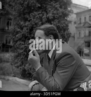 Dieses Archivfoto von 1975 ist ein intimes, ehrliches Profilporträt des blinden Kriegsveteranen Shutenko in Swiatohirsk (damals Slavyanogorsk), ukrainische SSR. Während des 30. Siegtages wird ein nachdenklicher, informeller Moment festgehalten. Shutenko sitzt in Anzug und Medaille auf einer Parkbank und raucht eine Zigarette in einem langen Halter. Im Hintergrund sind ein großes historisches Gebäude und Bäume sanft fokussiert. Stockfoto