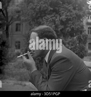 Dieses Archivfoto von 1975 ist ein intimes, ehrliches Profilporträt des blinden Kriegsveteranen Shutenko in Swiatohirsk (damals Slavyanogorsk), ukrainische SSR. Während des 30. Siegtages wird ein nachdenklicher, informeller Moment festgehalten. Shutenko sitzt in Anzug und Medaille auf einer Parkbank und raucht eine Zigarette in einem langen Halter. Im Hintergrund sind ein großes historisches Gebäude und Bäume sanft fokussiert. Stockfoto