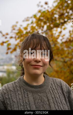 Lächelnde Frau im grauen Pullover draußen im Herbstpark, umgeben von gelben Blättern. Stockfoto
